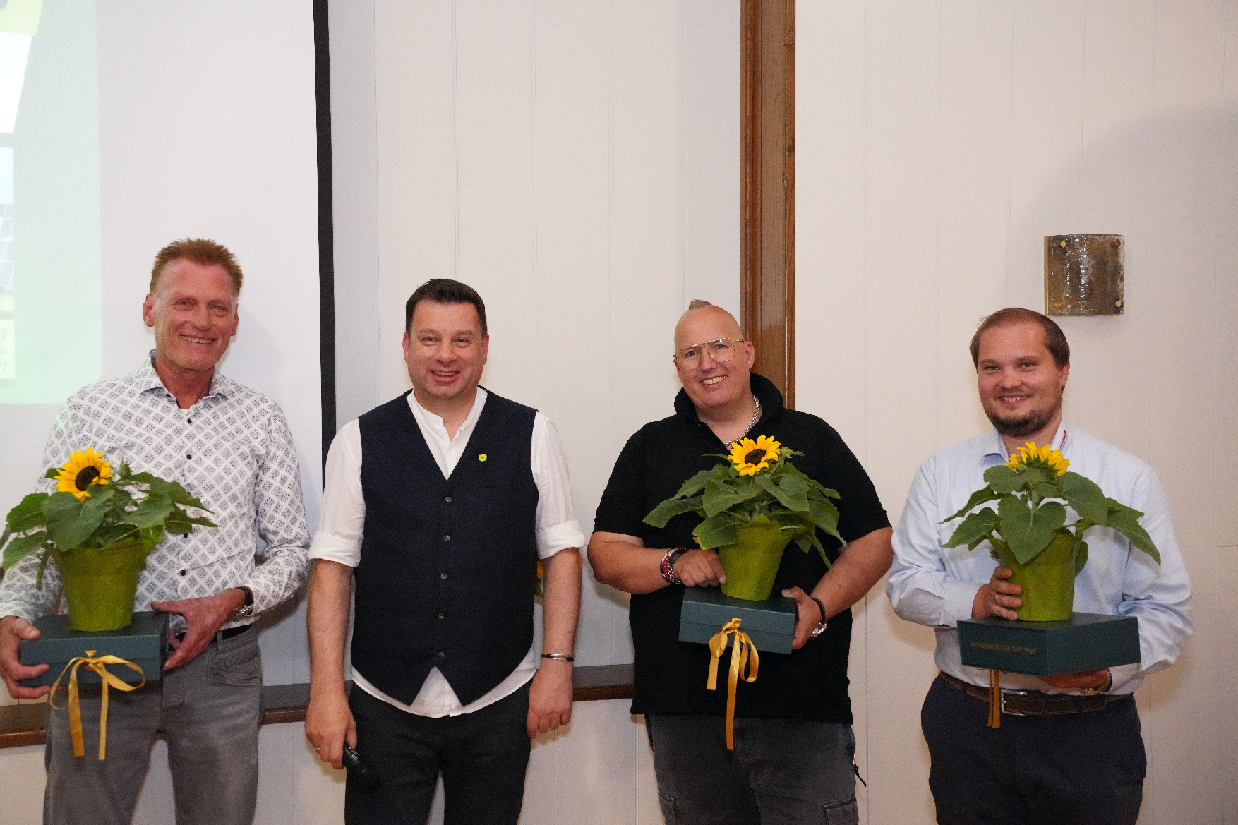 Vier Männer, drei davon mit Blumen, stehen lächelnd vor einer Wand.