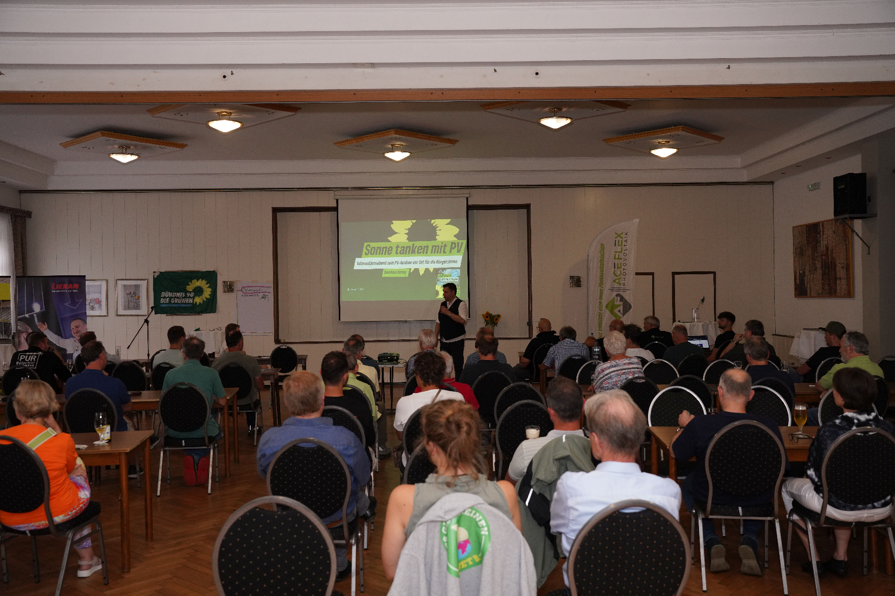 Ein Saal. Viele Menschen sitzen auf Stühlen. Ein Mann steht vorne. Hinter ihm auf der Leinwand steht groß in grün: "Sonne tanken mit PV".